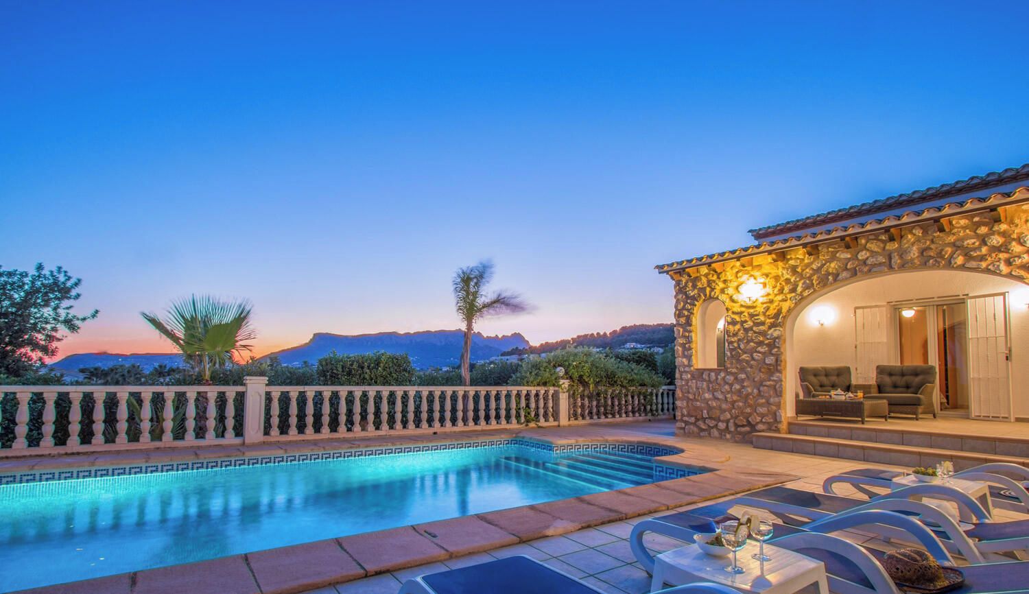 A pool area with a stone building, lounge chairs, and a view of the mountains.
