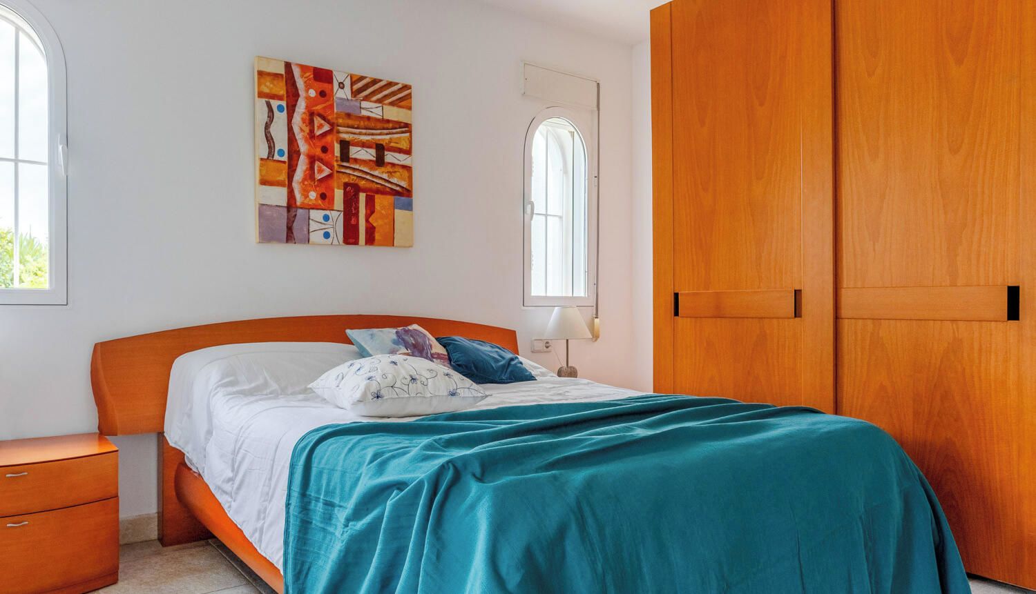 A bedroom with a bed, a nightstand, a wardrobe, a window, and a painting on the wall.