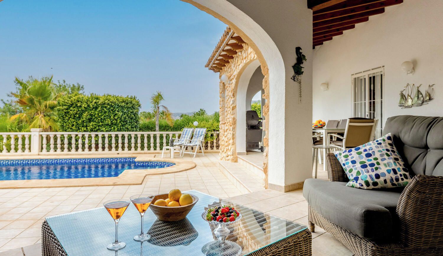 A poolside area with a glass table, lounge chairs, and a sofa with a patterned pillow.