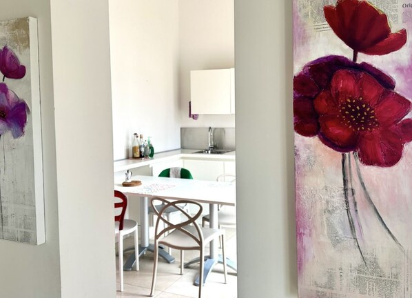 Un coin repas avec une table et des chaises blanches, une cuisine en arrière-plan et un tableau représentant des fleurs rouges accroché au mur.
