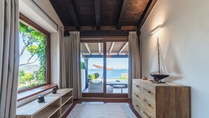 A room with a wooden ceiling, a large window with curtains, a wooden dresser, a small table, and a view of the sea through a glass door. - Villa Caterina, amazing front sea 2 Br villa with private beach (Punta Molara)