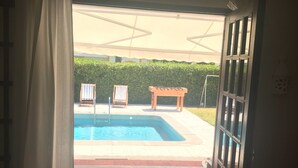 Una porta oberta que dona a una zona de piscina amb una tenda de sol, dues cadiretes i una taula de fusta.