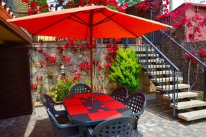 Um pátio externo com mesa, cadeiras e um guarda-sol vermelho, além de uma escadaria.