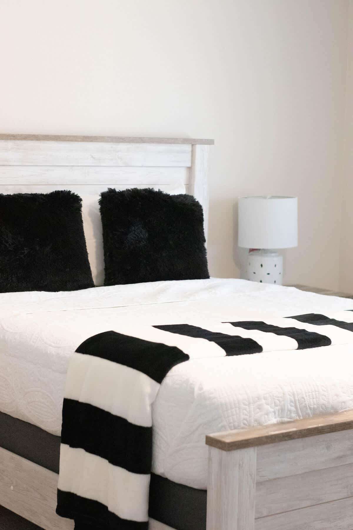 A bed with a white and black striped blanket, two black pillows, and a white bedside table with a lamp.