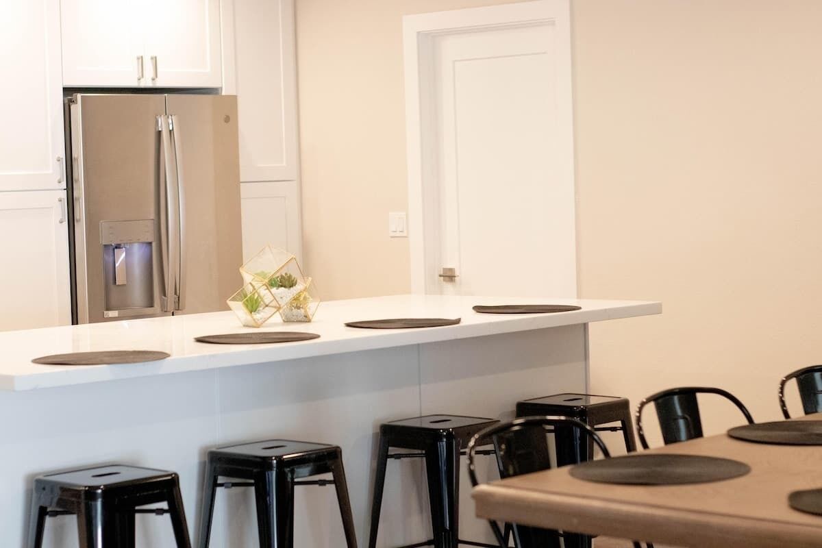 A modern kitchen with a stainless steel refrigerator, white cabinets, and a white countertop. There are black stools and a table with a round mat.