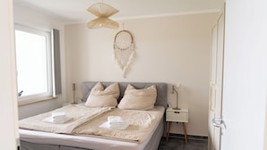 Una cama bien hecha con almohadas y una colcha, una mesita de noche con lámpara, y un atrapasueños en la pared.