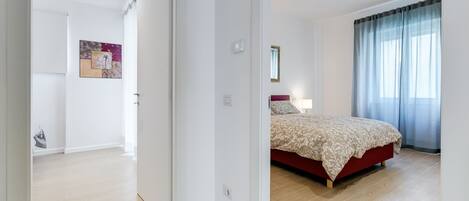 A bedroom with a bed, a nightstand, a window with curtains, and a door leading to another room.