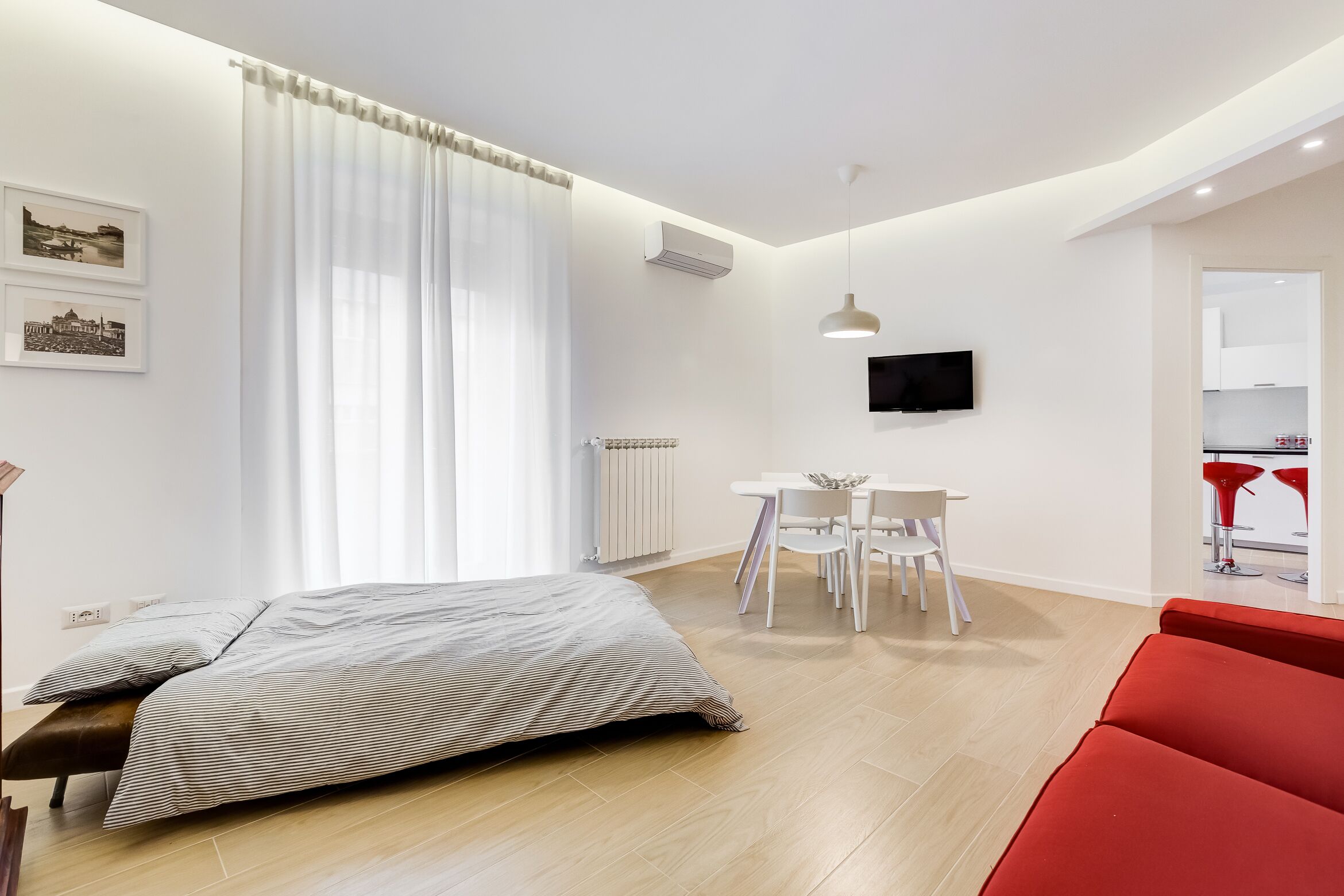 A modern hotel room with a bed, a dining area with a table and chairs, a TV mounted on the wall, and a red chair in the foreground.