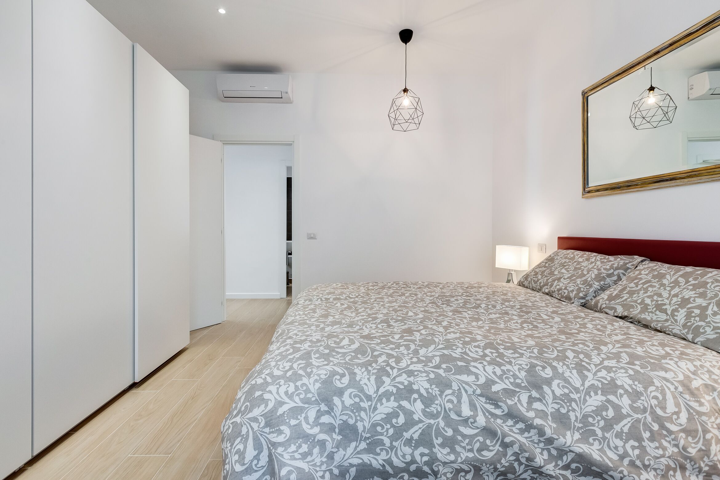 A bedroom with a large bed, a red headboard, a geometric pendant light, and a mirror on the wall.