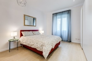 A bedroom with a red bed, a bedside table with a lamp, a mirror on the wall, and a window with curtains.