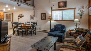Una sala de estar con sofá, una mesa de centro, televisor, mesa de comedor con sillas y una escalera.