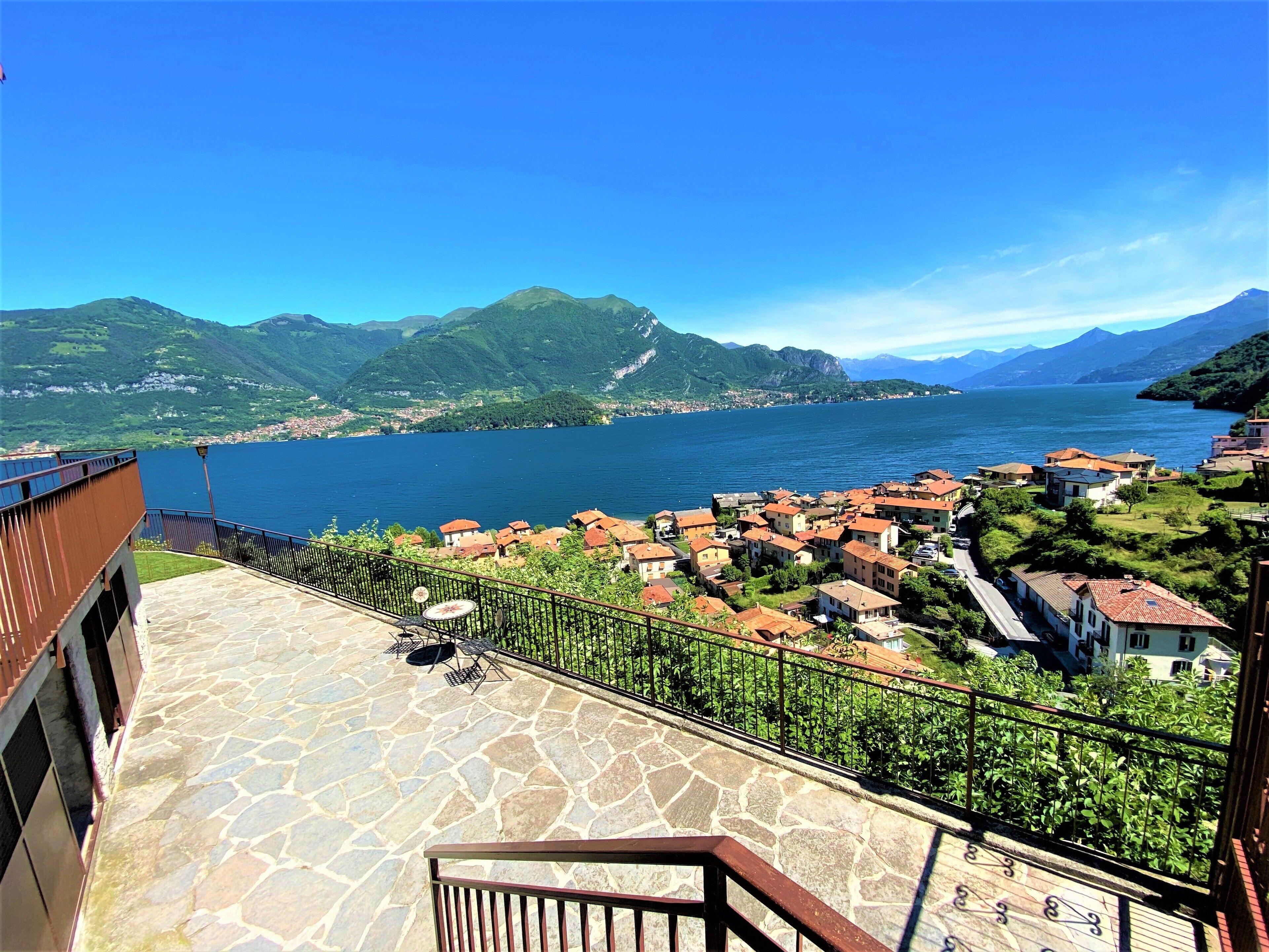 A terrace with stone flooring and a metal railing overlooking a lake with a village and mountains in the background.