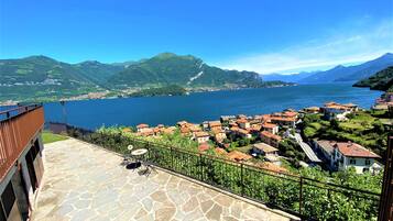 A terrace with stone flooring and a metal railing overlooking a lake with a village and mountains in the background.