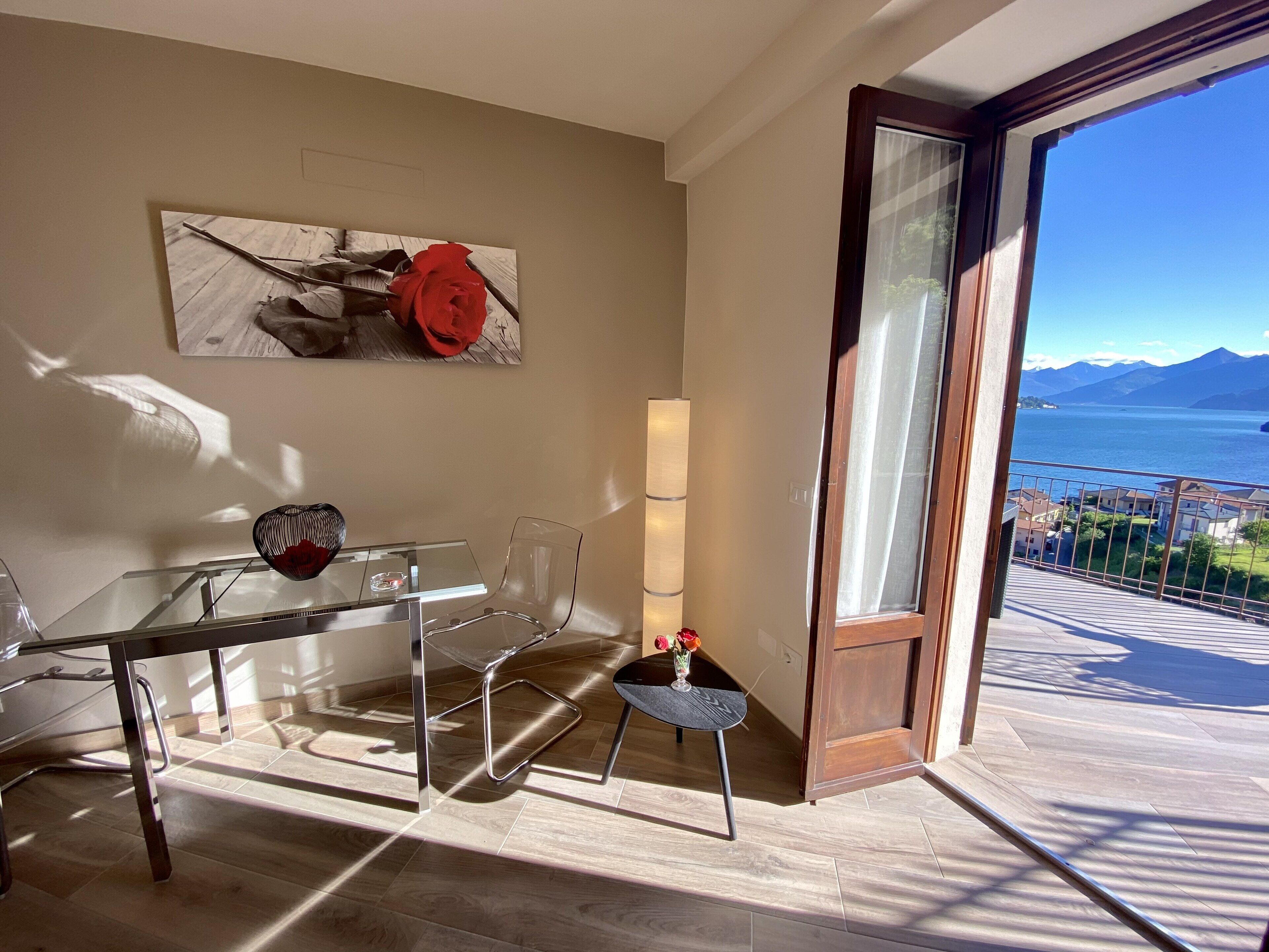 Un salon moderne avec une table en verre, des chaises transparentes, un vase rempli de fleurs et un tableau accroché au mur. Une grande fenêtre offre une vue sur un lac et des montagnes.