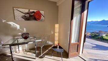 Un salon moderne avec une table en verre, des chaises transparentes, un vase rempli de fleurs et un tableau accroché au mur. Une grande fenêtre offre une vue sur un lac et des montagnes.
