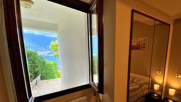 A room with a view of a mountainous landscape through a window, a bed, a bedside table with a lamp, and a mirror on the wall.