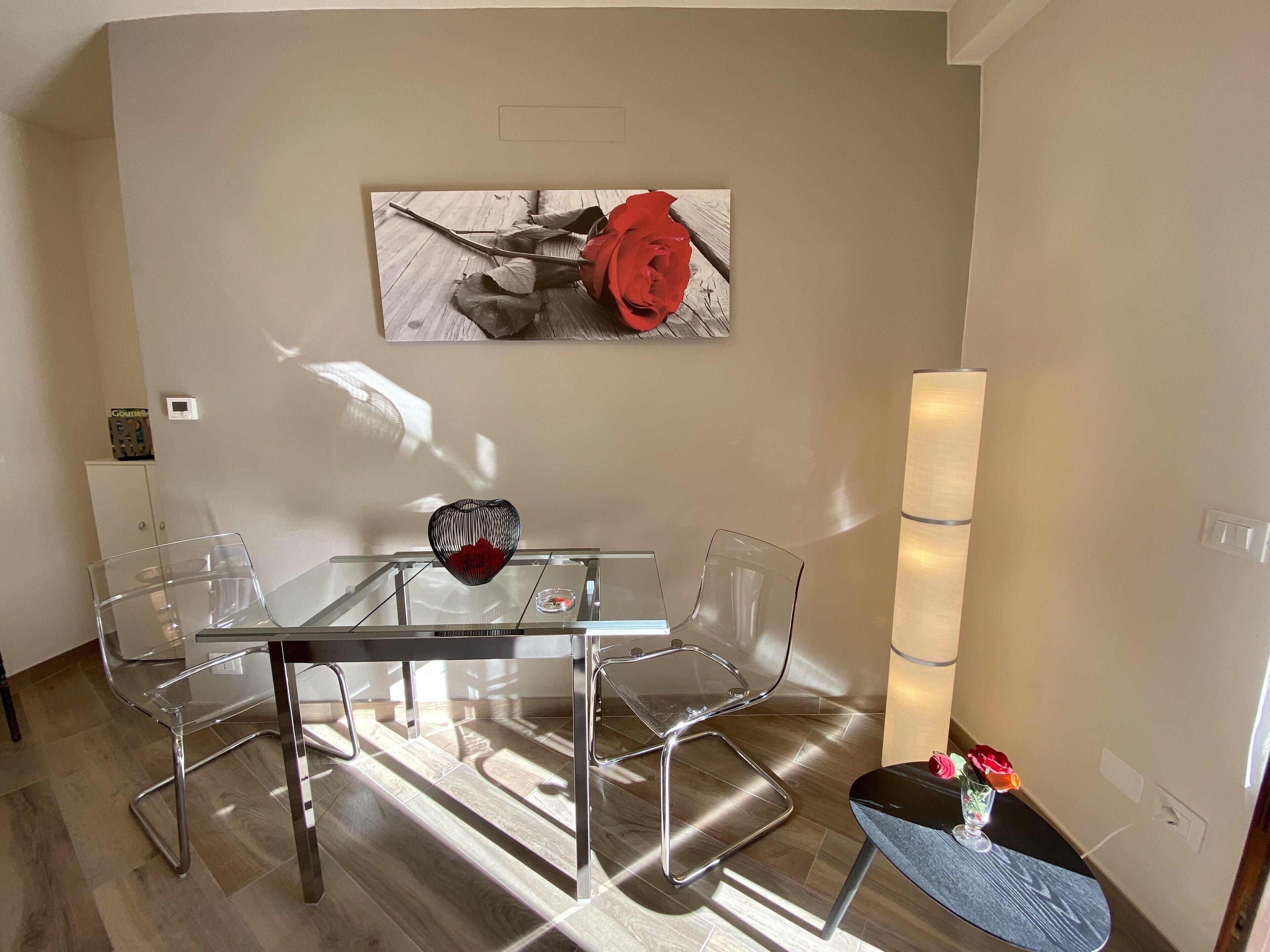 A modern dining area with a glass table and metal chairs, a decorative vase with a red flower, and a wall-mounted artwork.