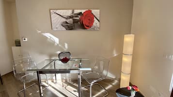 A modern dining area with a glass table and metal chairs, a decorative vase with a red flower, and a wall-mounted artwork.