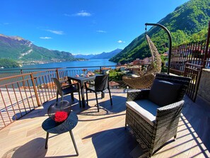 Eine Terrasse mit Korbmöbeln, einem Tisch mit Tassen und einer Vase, und Blick auf einen See und Berge.