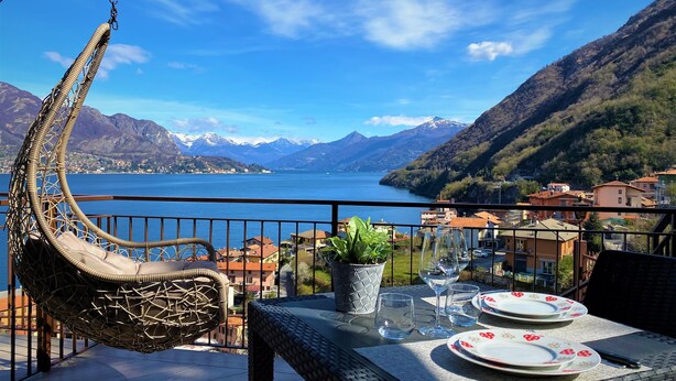 A balcony with a wicker chair, a table set with plates and glasses, and a view of a lake with mountains in the background. - RUBINO NEW!  NEAR BELLAGIO! BALCONY GARDEN, &  STUNNING VIEW! (lezzeno)