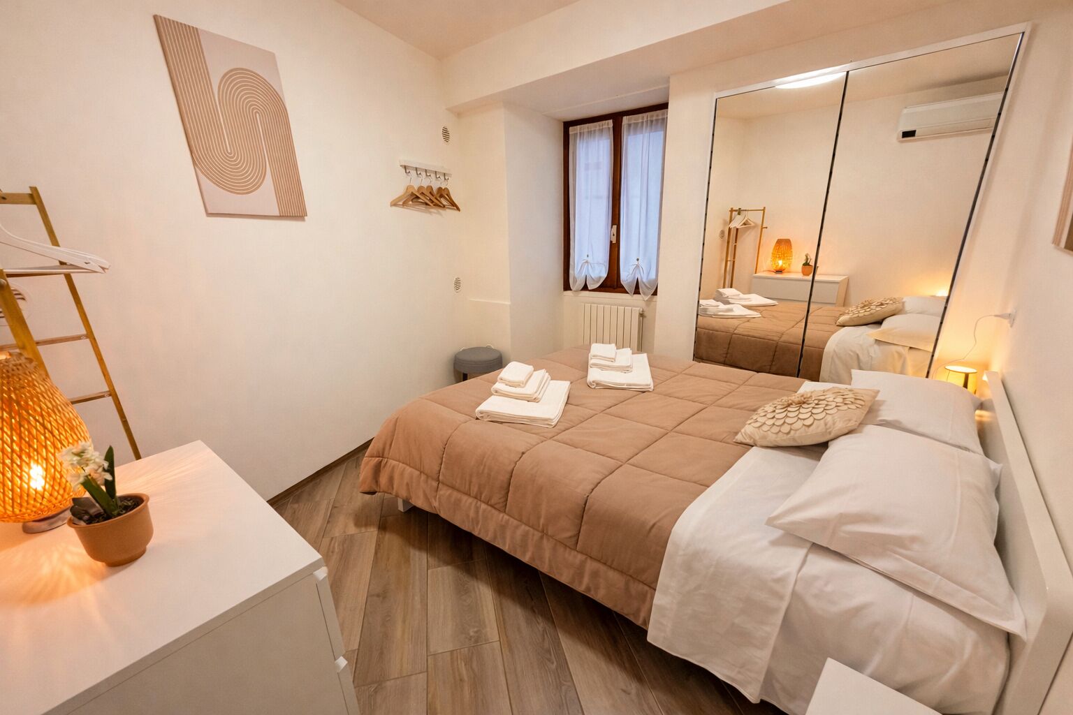 Quarto com cama, mesa de cabeceira, espelho, janela com cortinas e uma obra de arte na parede.