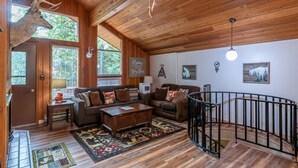 A wooden interior with a deer head mounted on the wall, a living room with sofas and a coffee table, and a staircase with a railing. - Escape to Ptarmigan Village 79 (Whitefish)