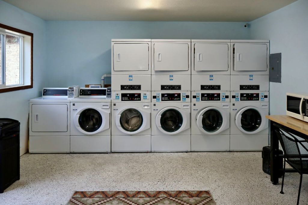 A laundry room with multiple washing machines and dryers, a small table with a microwave, and a window providing natural light.