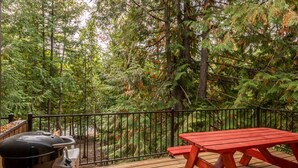 A deck with a barbecue grill and a red picnic table, surrounded by dense forest. - Escape to Ptarmigan Village 79 (Whitefish)