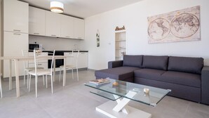 A modern living room with a glass coffee table, grey sofa, and white dining chairs. - Luxury Seafront Dependance in Fertilia (Fertilia)