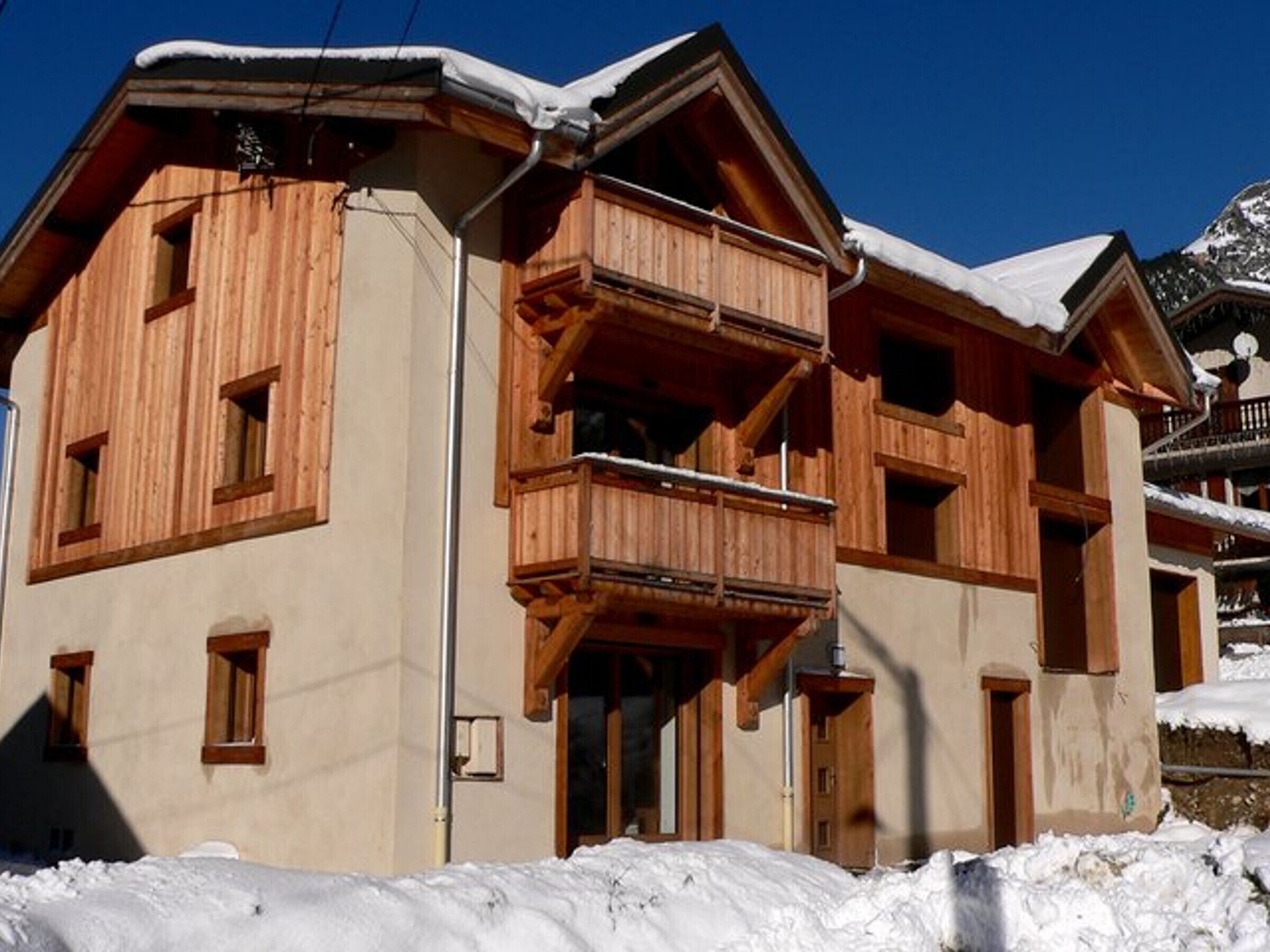 A wooden chalet with a balcony, a snow-covered roof, and a mountain backdrop.