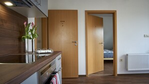 A modern hotel room with a wooden door labeled "KÜCHE + 2", a bed, a radiator, and a bathroom with a towel rack. - Apartment Noerdliches Muensterland (Hopsten)