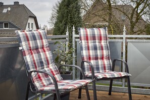Deux chaises de patio destinées à être utilisées à l’extérieur, dotées de coussins à carreaux, une clôture en métal et un pot de fleurs.