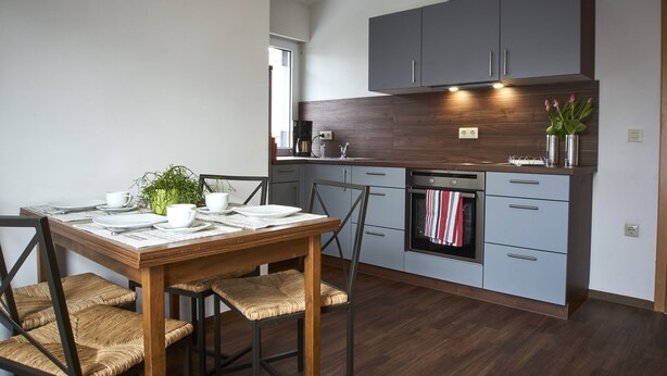 A modern kitchen with grey cabinets, a wooden dining table, and a built-in oven. - Apartment Noerdliches Muensterland (Hopsten)