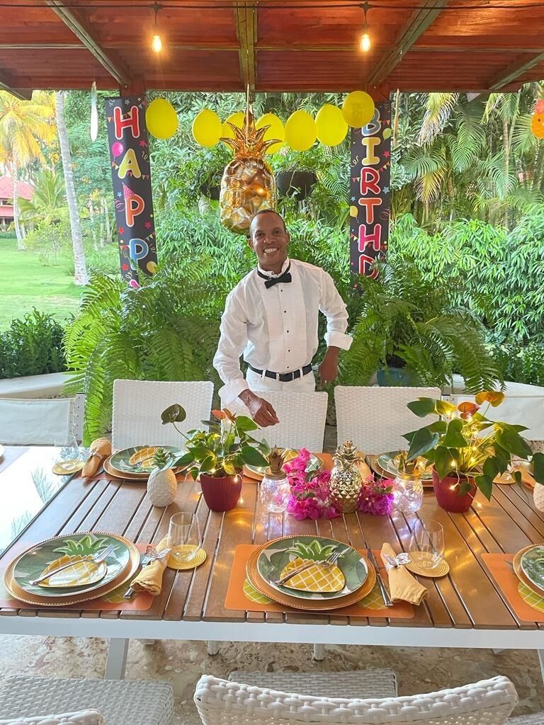 A table set outdoors with a tropical backdrop, a man standing behind it, and a 'HAPPY BIRTHDAY' banner.