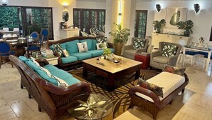 A spacious living room with a sofa set, coffee table, and chairs. There is a dining area with a table and chairs. The room has large windows and a view of greenery outside. - 120 +5 STAR reviews below 3 Min to Beach English speaking Chef  Butler Meal Plan (La Romana)