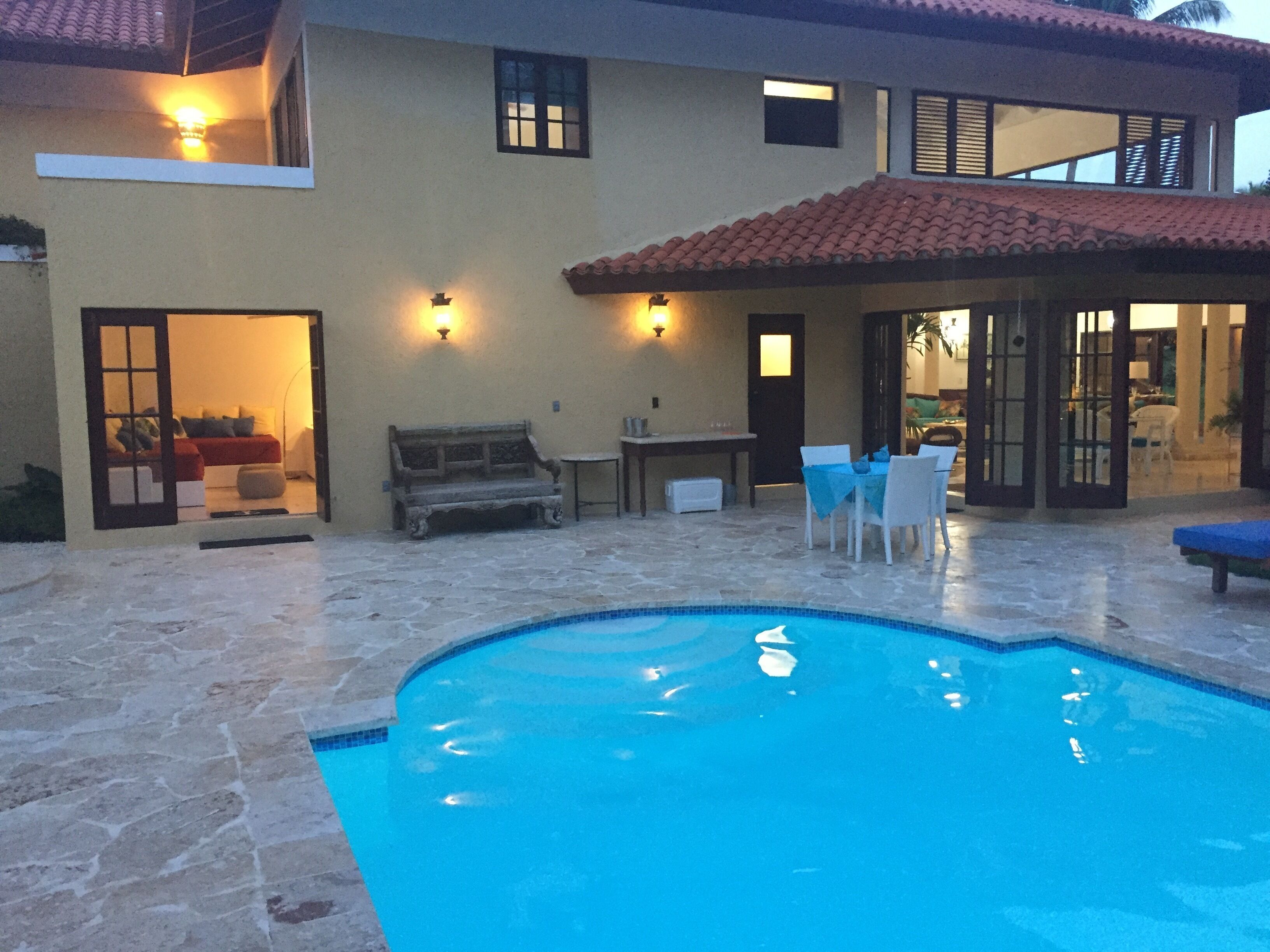 A swimming pool with stone tiles, a patio area with a bench, and a dining table with chairs.