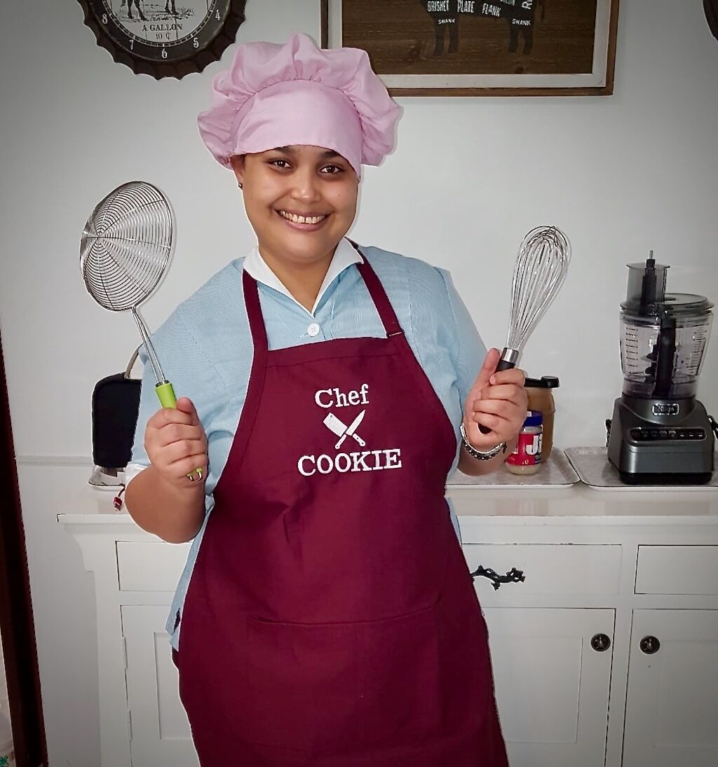 A person wearing a chef's hat and apron, holding a whisk and a spatula.