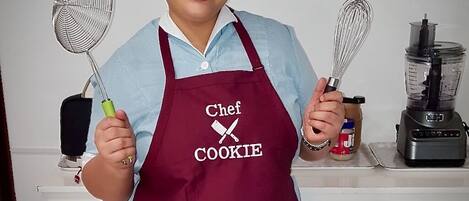 A person wearing a chef's hat and apron, holding a whisk and a spatula.