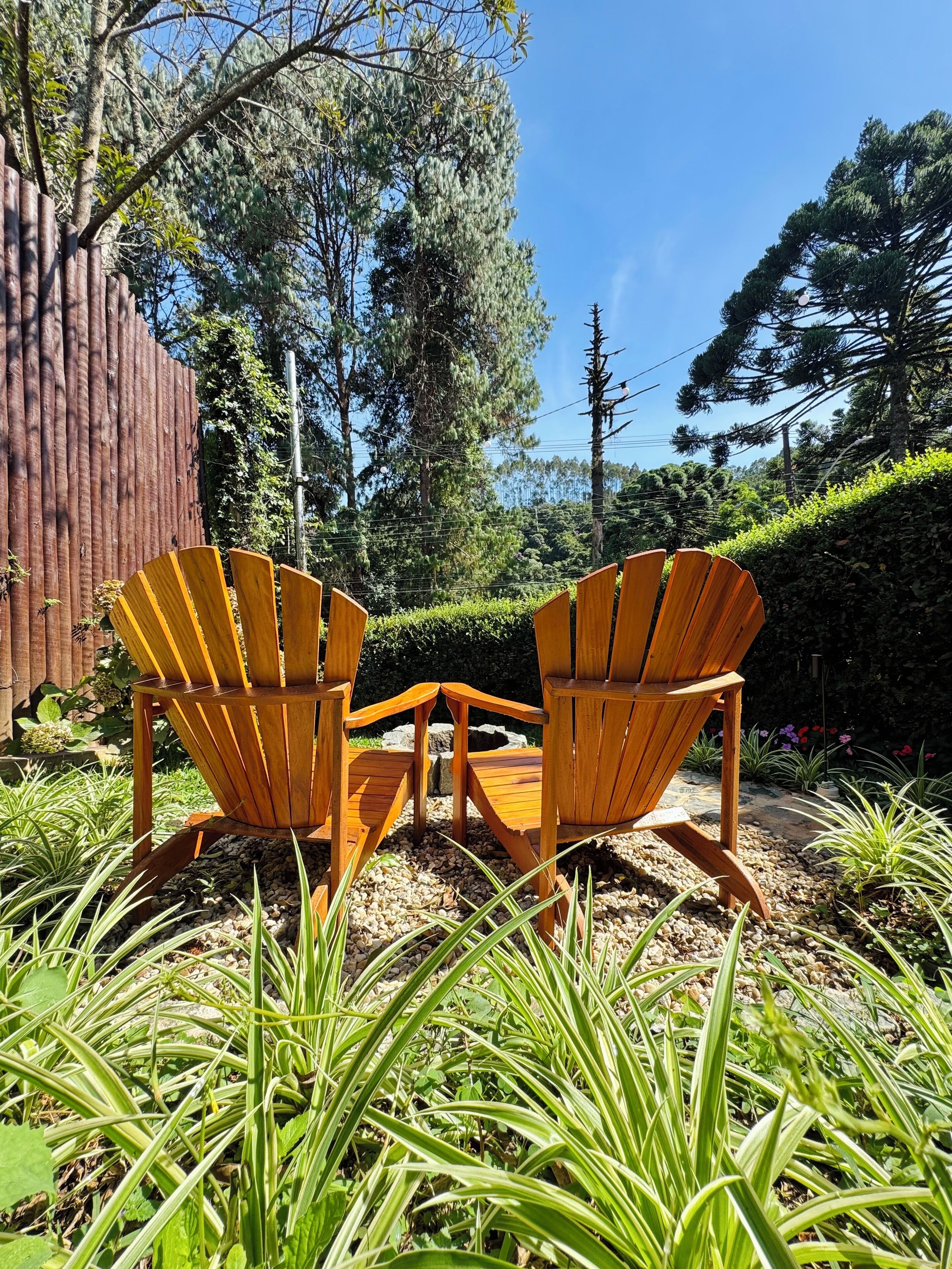 Two Adirondack chairs in a garden setting with a wooden fence, greenery, and trees in the background.
