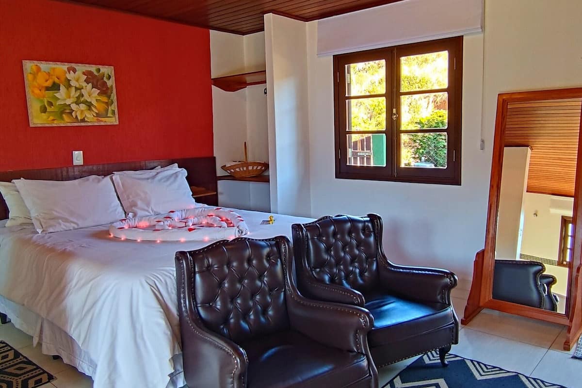 A bedroom with a bed, two leather chairs, a patterned rug, a wooden ceiling, and a window with a view.