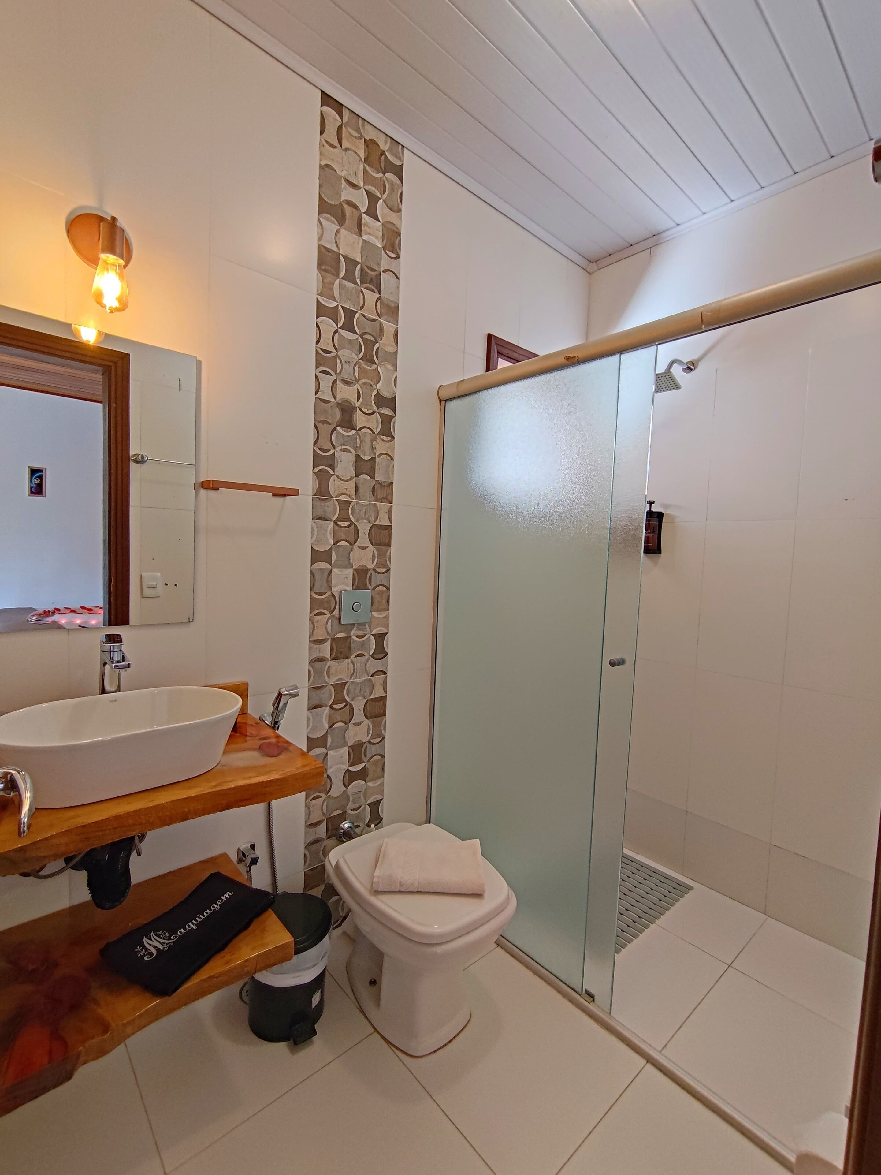 Casa de banho com lavatório branco, espelho e sanita, cabine de duche em vidro e parede com padrão.