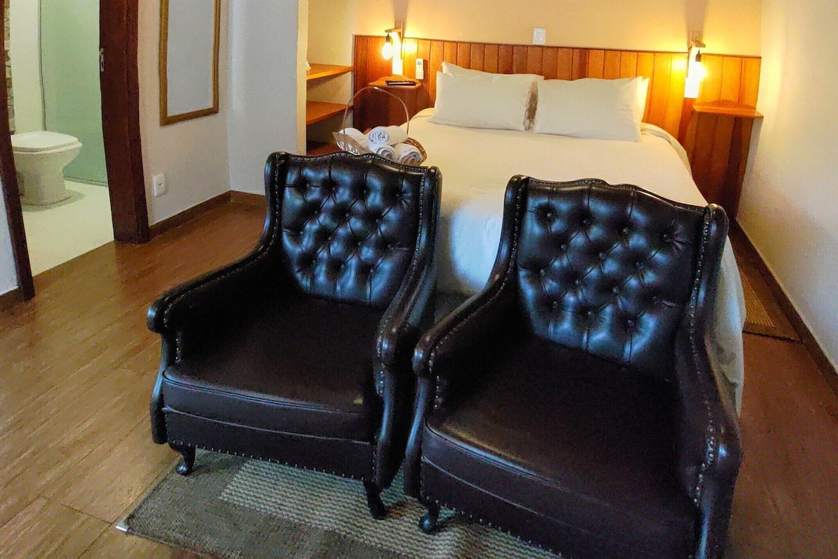 A hotel room with a wooden ceiling, a bed with two pillows, two bedside lamps, and two black leather chairs.