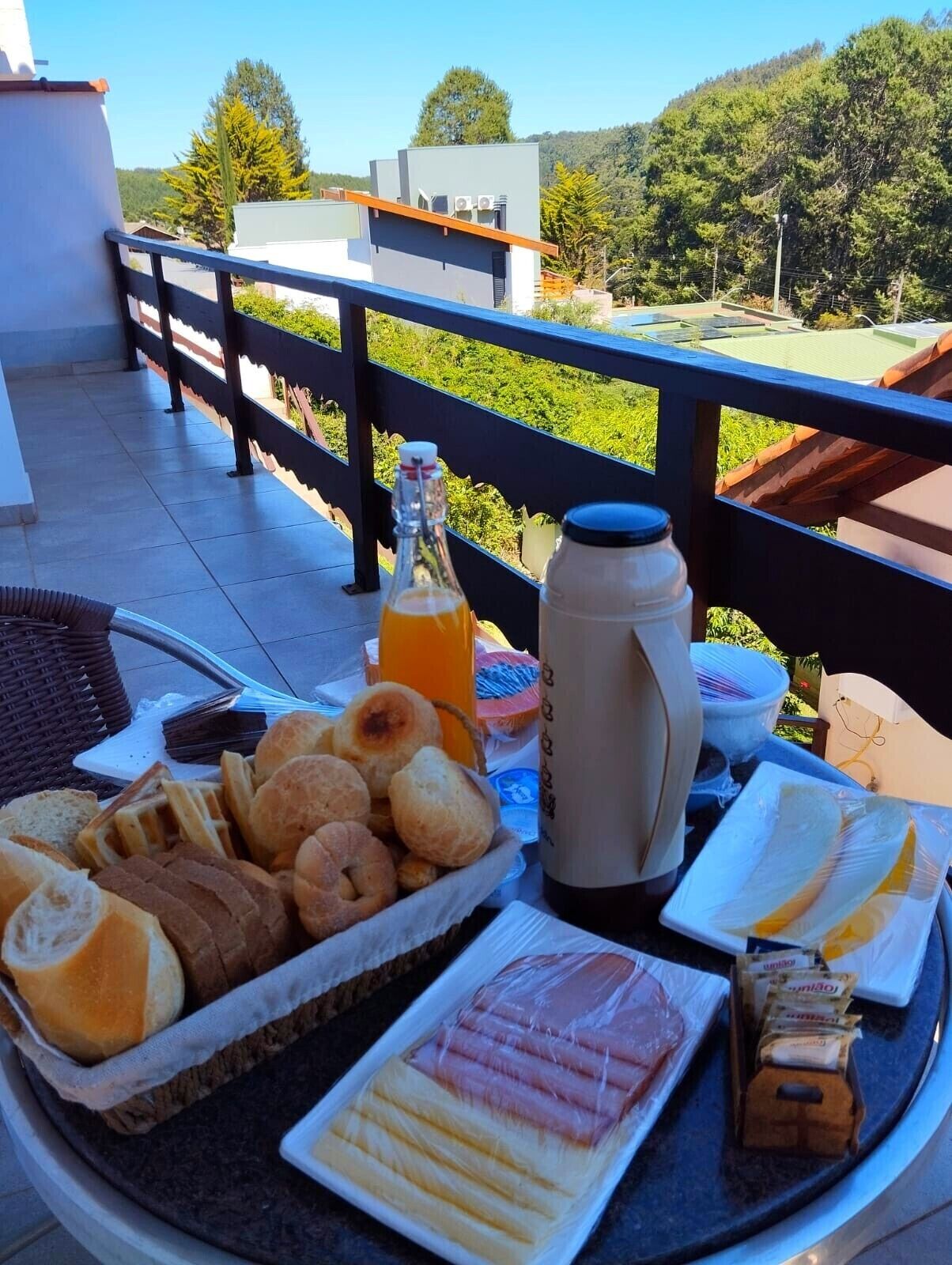 Ein Frühstückstisch auf einem Balkon mit Brot, Gebäck, einer Orangensaftflasche, einem Thermosbehälter und einer Platte Schinken.