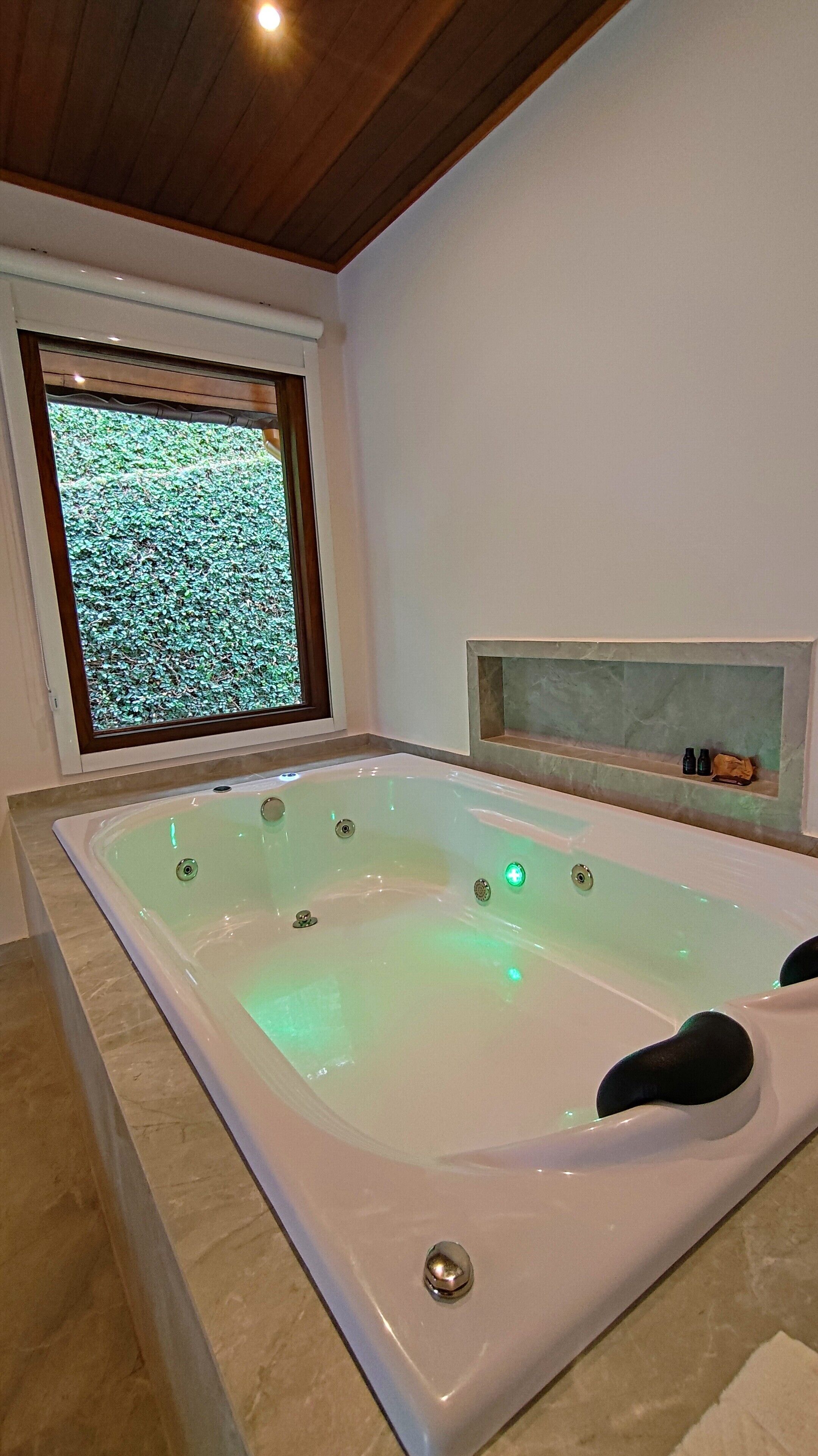 A modern bathroom with a large bathtub, a window with a green leafy view, and a built-in shelf with toiletries.