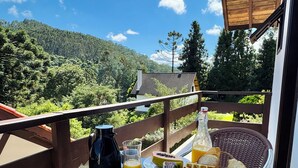 Balcón con mesa lista para el desayuno, que incluye pan, bollería, fruta y bebidas, con vistas a un paisaje boscoso.