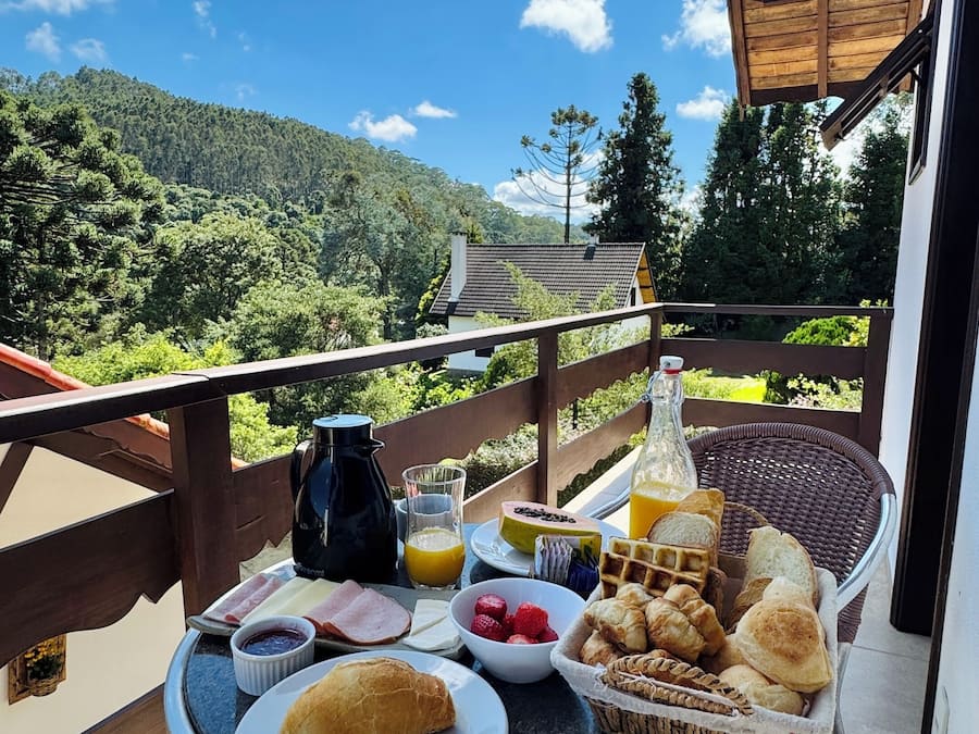 Un balcon avec une table dressée pour le petit-déjeuner, comprenant du pain, des viennoiseries, des fruits et des boissons, donnant sur un paysage boisé.