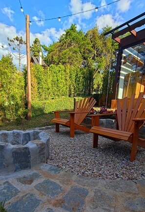 Um deck de madeira com cadeiras Adirondack, rodeado por uma borda de pedra e pedrinhas, com um jardim e árvores ao fundo.