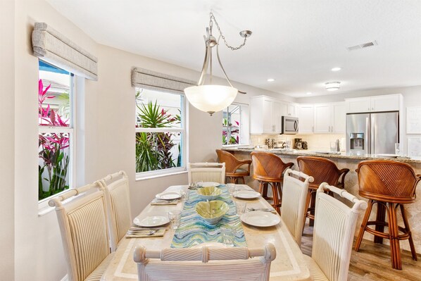 A dining area with a table set for four, surrounded by wicker chairs, a hanging light fixture, and a view of a kitchen with white cabinets and stainless steel appliances.