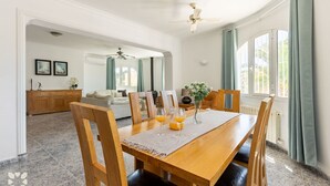 A dining area with a wooden table, chairs, and a ceiling fan. There is a window with curtains and a view of greenery outside. - Villa MACKAY by Abahana Villas (Calpe)