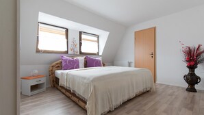 Un dormitorio con una cama, dos ventanas, una mesita de noche, un aparador y un florero con flores.